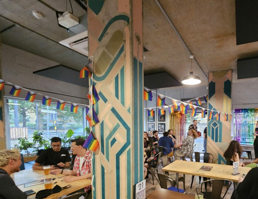 Magic The Gay-thering event in Bad Moon café London. Pride flag bunting is draped across the room with people sat playing MTG. 